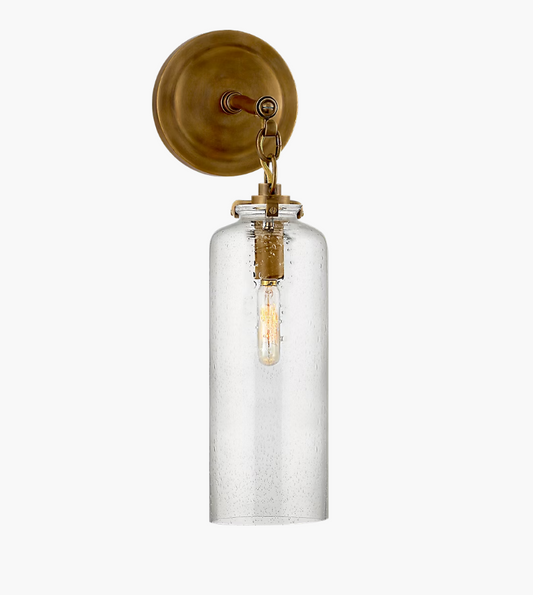 Wall-mounted light fixture with clear glass cylindrical shade and brass metal backplate.