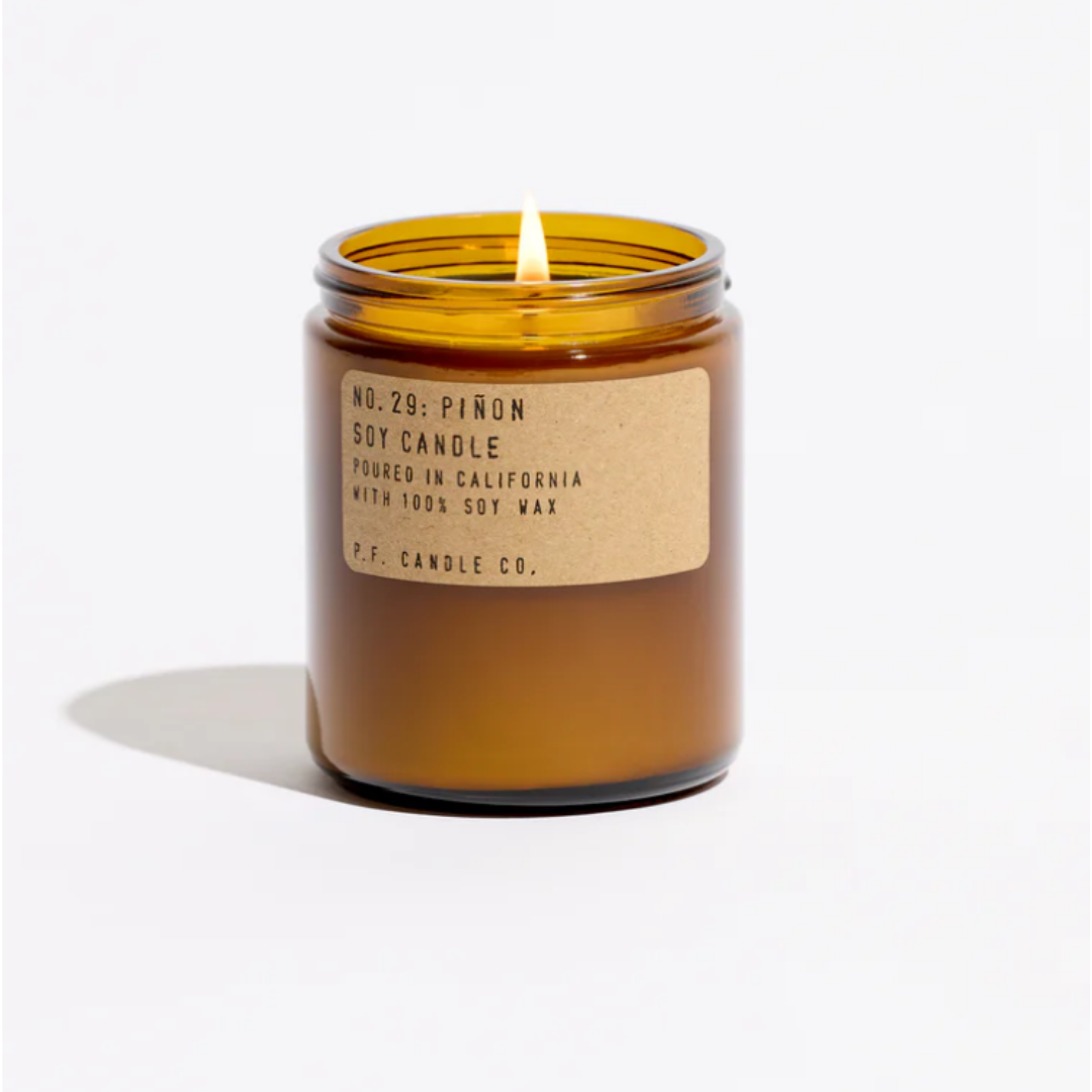 Amber glass candle jar labeled “P.F. Candle Co. Piñon” with a lit wick on a white background.
